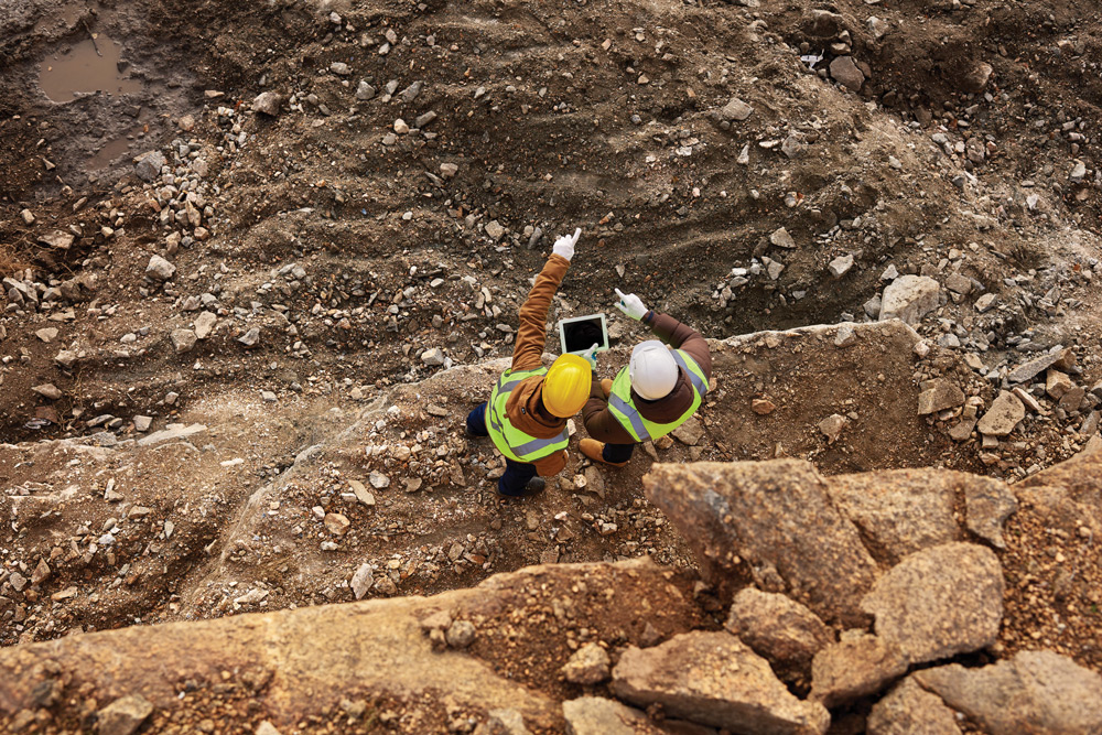 Overhead shot of two men at a construction site in high-visibility protective gear looking at plans on a tablet