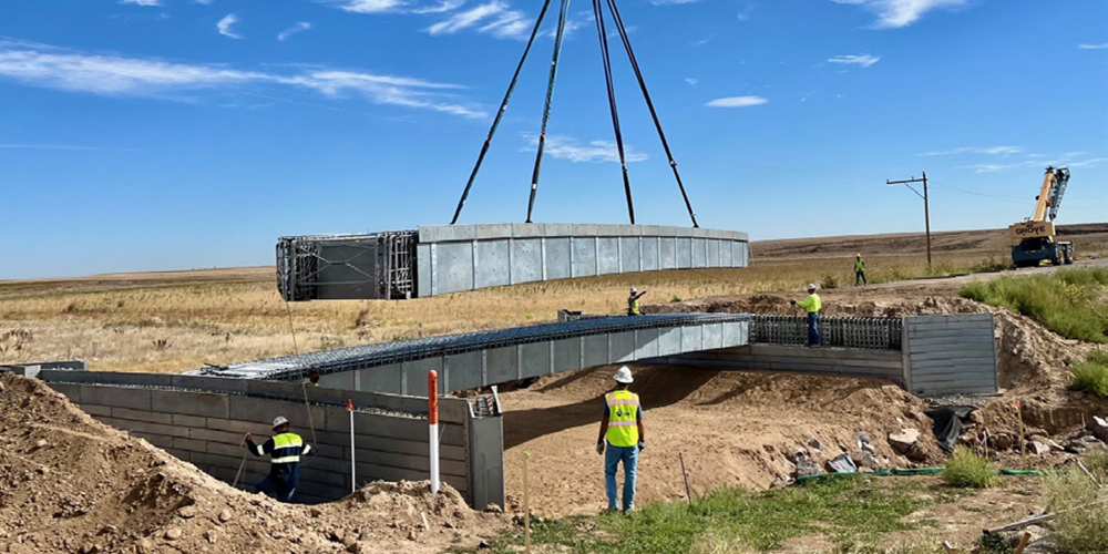 Bridge deck being lowered into place by crane with construction workers in foreground and background.