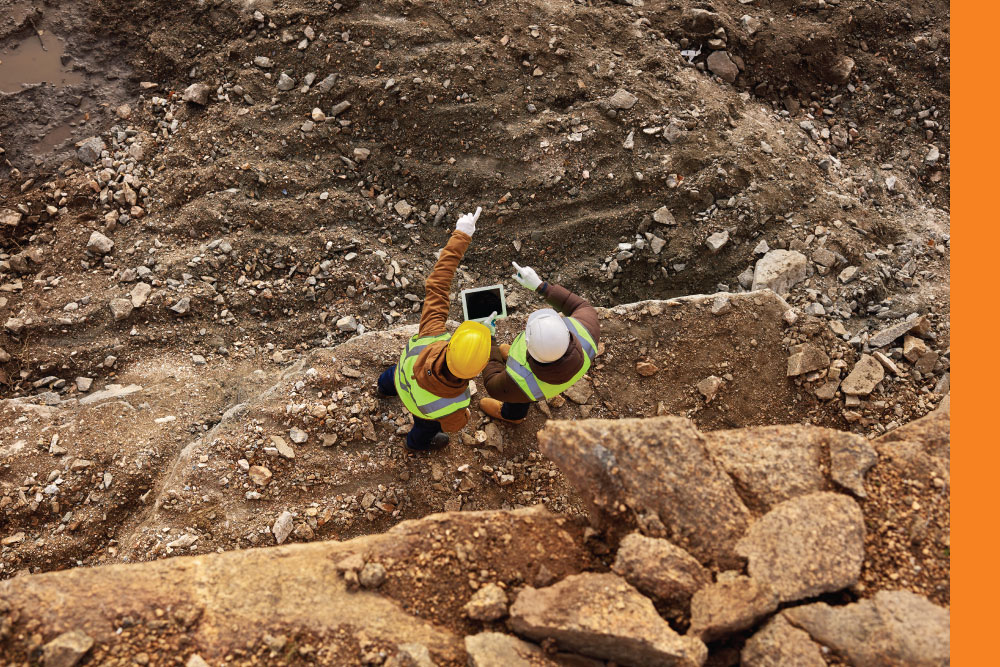 Two construction workers in high visibility gear and hard hats looking over a construction site (view from above).